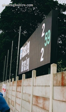 A large digital scoreboard at an outdoor sports venue displays a score of 2-2. Spectators are gathered, observing the event with interest. The scene is enclosed by a tall metal fence and surrounded by lush green trees, indicating a natural setting. Overcast skies add a muted tone to the environment.