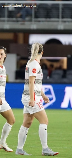 Soccer players in team uniforms stand on a grass field with a large digital advertisement screen displaying the acronym 'NWSL' in the background. The setting appears to be a stadium with rows of empty seats visible above the screen.