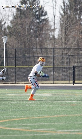 A group of athletes engaged in a sports game on a green field. One player is running forward with possession of the ball, wearing colorful socks and a checkered uniform, while another player in dark clothing dives to intercept or tackle. Other players are present in the background. Leafless trees and a metal fence surround the field.