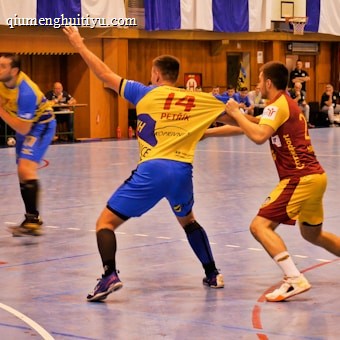A dynamic indoor sports scene with players in motion on a court. Two athletes in blue and yellow uniforms are engaged in an intense handball game, with one player gripping the other's jersey. The background shows other players, several seated people, and advertisements on the walls.