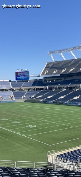 A large, empty sports stadium with blue seats surrounding a well-maintained green grass field. Bright sunlight creates clear shadows, and there are two goal posts on opposite ends of the field. A big electronic scoreboard displays some graphics, and the sky is clear and blue.